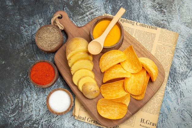 Free photo above view of tasty homemade chips cut potato slices on wooden cutting board and different spices on newspaper on gray table