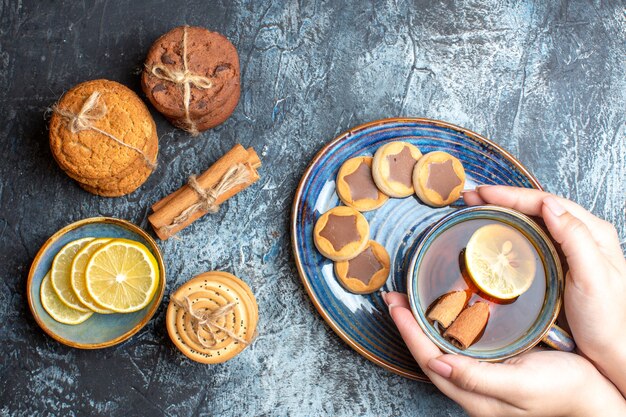 Above view of tea time with various cookies and hand holding a cup of black tea with cinnamon on a blue tray on dark background