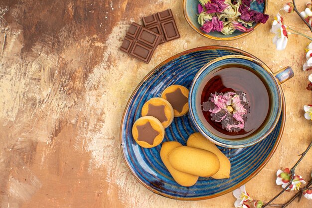 Above view of various biscuits a cup of tea and flowers chocolate bars on mixed color table