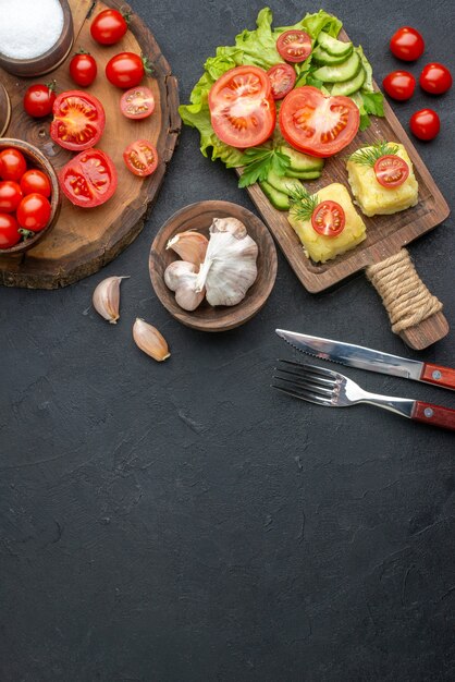 Above view of whole cut fresh vegetables and spices on wooden board white towel cutlery set cheese on black surface