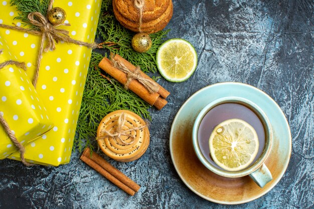 Above view of Xmas background with beautiful yellow gift boxes and stacked cookies lemon cinnamon limes next to a cup of black tea on dark background