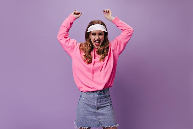 Free photo vigorous woman in pink hoodie and cap shouts on purple background emotional girl in denim skirt dancing on isolated backdrop