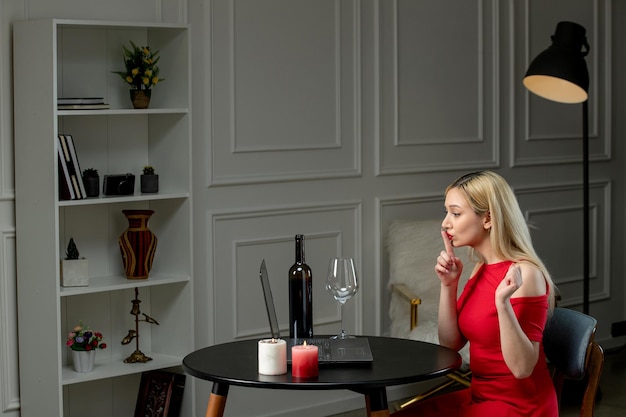 Free photo virtual love cute blonde girl in red dress on distance date with wine candles showing silence sign