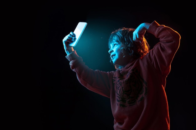 Vlogging with smartphone. Caucasian boy's portrait on dark studio background in neon light. Beautiful curly-haired model.