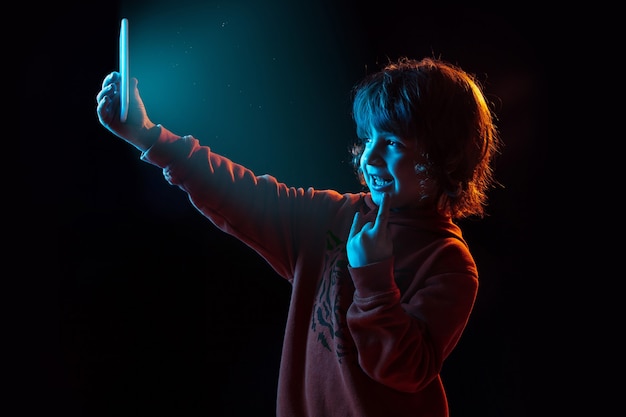 Vlogging with smartphone. Caucasian boy's portrait on dark  wall in neon light. Beautiful curly-haired model. Concept of human emotions, facial expression, sales, ad, modern tech, gadgets.