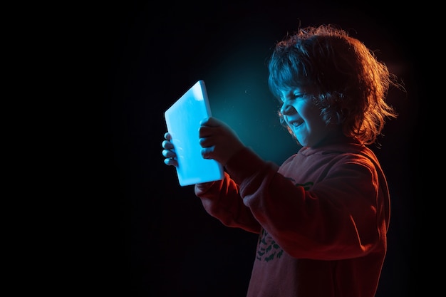 Vlogging with tablet, playing. Caucasian boy's portrait on dark  wall in neon light. Beautiful curly model. Concept of human emotions, facial expression, sales, ad, modern tech, gadgets.