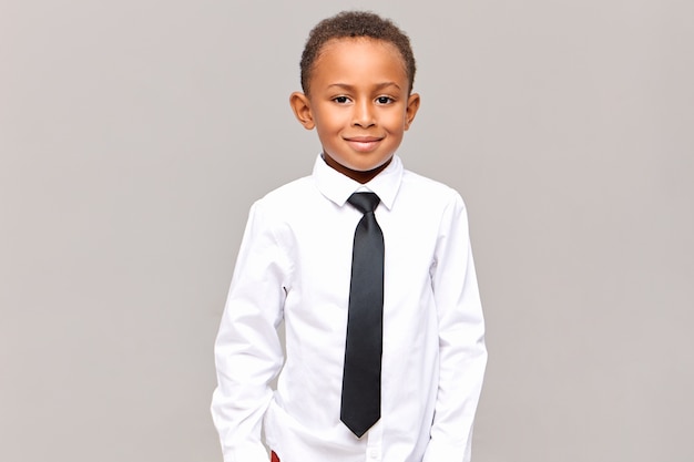 Waist up  handsome neat dark skinned male elementary pupil posing isolated dressed in white clean ironed shirt and black elegant tie, ready for going to school, smiling