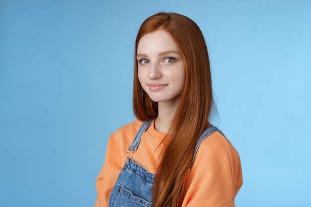 Waist-up kind sincere tender lovely redhead girl wearing orange shirt denim overalls standing half-turned smiling silly gentle grin camera looking friendly pleasantly walk alone blue background.