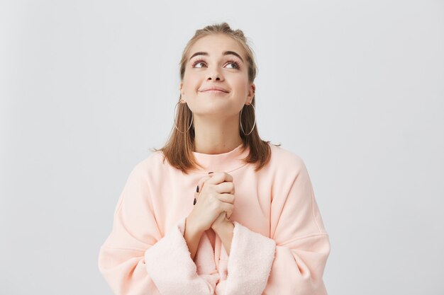 Waist up portrait of cheerful caucasian female with fair hair posing in studio with looking upwards. Girl dressed in pink sweatshirt keeping palms pressed together, having begging look.