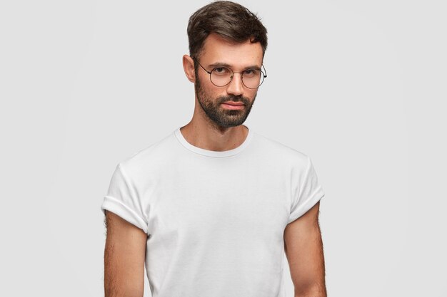 Waist up portrait of handsome bearded male with serious expression, contemplates about something, wears round spectacles and white casual t-shirt, poses indoor. People and facial expressions