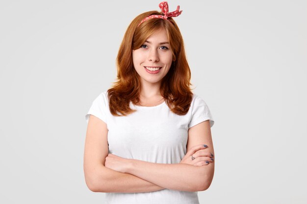 Waist up portrait of happy young female with broad smile, appealing appearance, keeps hands crossed, wears retro style clothes, happy to hear good news, isolated on white studio