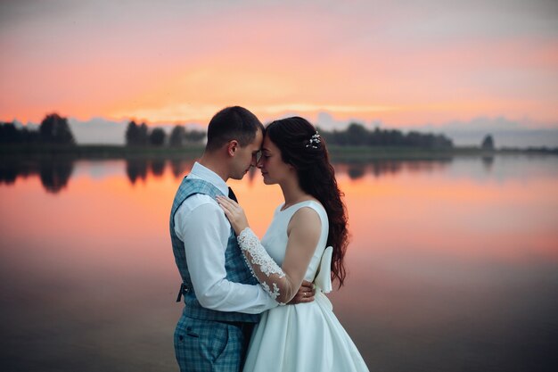 Waist up of romantic wedding couple hugging and posing by the lake at sunset with amazing view. Wedding couple in love concept