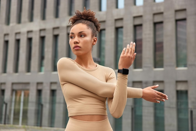 Free photo waist up shot of active slim woman stretches arms before workout dressed in top has curly combed hair concentrated away poses against blurred building background prepares for cardio training