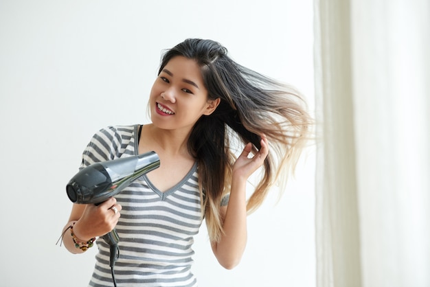 Waist up shot of Asian girl blow-drying her hair at home