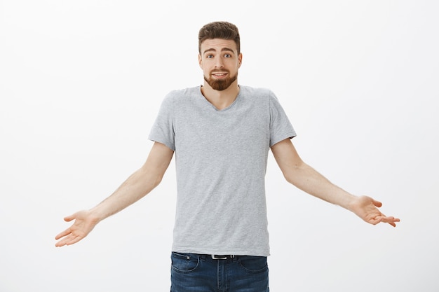 Waist-up shot of carefree clueless and unconcerned cute man with blue eyes and beard shrugging with hands spread aside and silly unaware smile being indifferent to what happening against gray wall