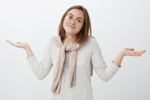 Free photo waist-up shot of clueless carefree uninvolved attractive european girl in glasses shrugging with hands spread aside smiling tilting head having no clue being unaware and careless over gray wall