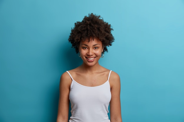 Free photo waist up shot of dark skinned young woman with afro hairstyle, smiles pleasantly, dressed in casual white vest, has pleasant talk, isolated on blue wall, expresses happiness and tenderness