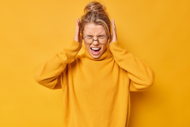 Waist up shot of emotional young blonde woman screams loudly covers ears with hands doesnt want to hear bothering sound wears spectacles and sweater isolated over yellow background Stop this