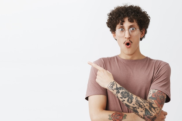 Free photo waist-up shot of excited and impressed emotive attractive male student with stylish moustache curly dark hair and tattooed arm gasping folding lips from amazement and pointing at upper left corner