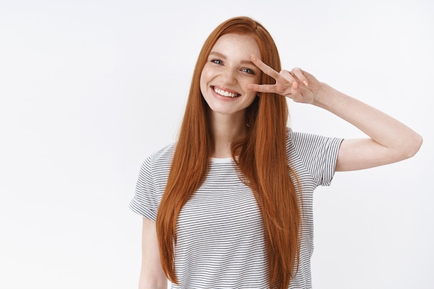 Free Photo waist-up shot friendly kind optimistic attractive young lucky teenage redhead girl blue eyes tilting head smiling white teeth show victory peace gesture enjoy party, standing white wall