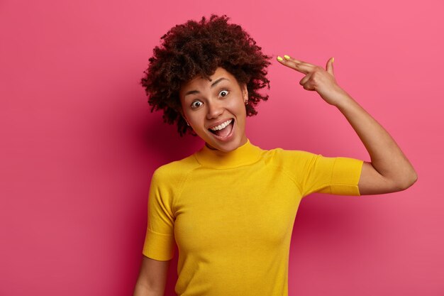 Waist up shot of funny Afro American woman makes finger gun pistol in temple, tilts head, laughs positively, wears yellow t shirt, pretends shooting and killing herself, isolated on pink wall