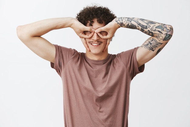 Free photo waist-up shot of funny and playful creative young curly-haired male with tattooed arm and moustache making eye mask with fingers smiling broadly, fooling around and having fun over gray wall