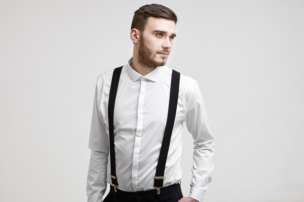 Waist up shot of good looking trendy guy with stylish beard standing at white studio wall having dreamy positive look, dressed in elegant white shirt with suspenders. People and lifestyle concept