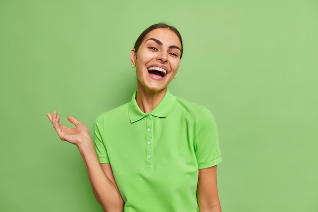 Free photo waist up shot of happy brunette woman dressed in casual t shirt smiles sincerely keeps arm raised laughs at funny anecdote wears casual t shirt isolated over green background. happiness concept