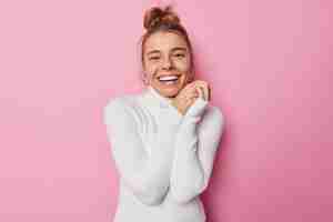 Free photo waist up shot of happy delighted european woman smiles broadly keeps hhands together feels glad to spend free time in company of friends wears white turtleneck poses against pink background.