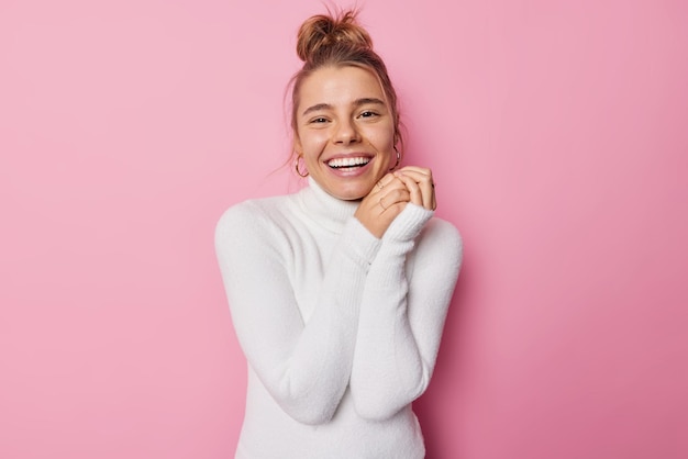 Waist up shot of happy delighted European woman smiles broadly keeps hhands together feels glad to spend free time in company of friends wears white turtleneck poses against pink background.