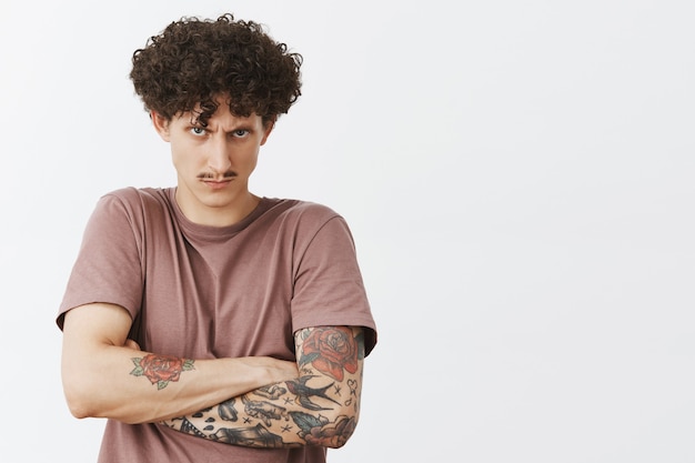 Waist-up shot of intense angry and pissed young curly-haired man with moustache and tattoos holding offence inside frowning looking from under forehead with scorn being mad over gray wall