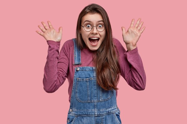 Waist up shot of joyful pretty girl shows palms, looks with overjoyed expression, wears casual clothes, round spectacles