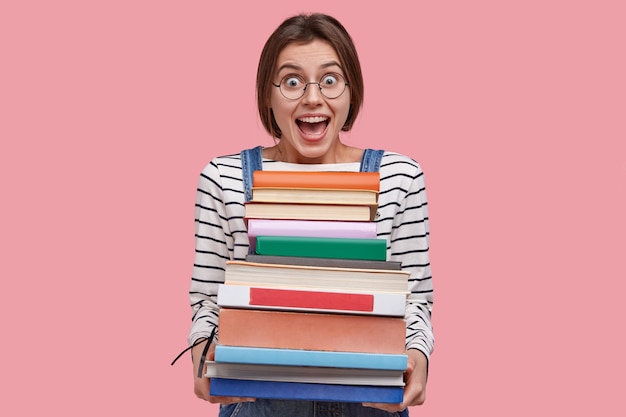 Free Photo waist up shot of joyful student in round spectacles, holds pile of books, prepares for seminar or writing report