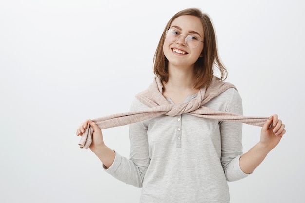 Free photo waist-up shot of playful attractive caucasian female brunette with trendy eyeglasses tying pullover over neck and smiling joyfully enjoying interesting conversation while hanging around with friends