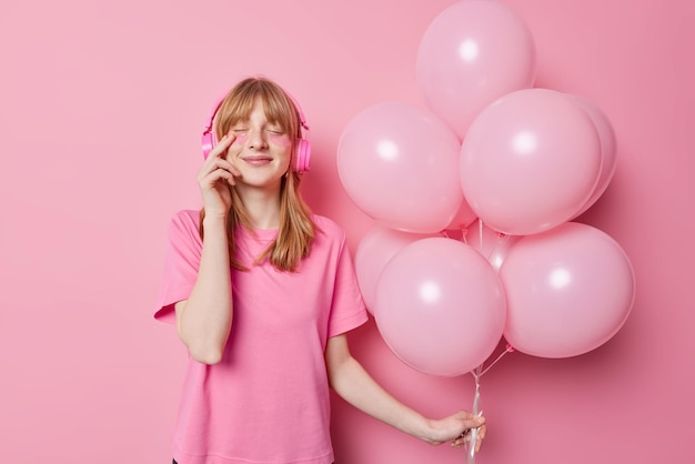 Free photo waist up shot of redhaired girl applies beauty patches for skin treatment keeps eyes closed listens music via headphones holds bunch of balloons enjoys birthday celebration isolated on pink wall