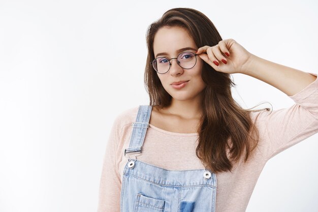 Waist-up shot of sensual and flirty good-looking female student with brown hair touching frame of glasses folding lips cute making romantic gazes at front standing seductive over gray wall.