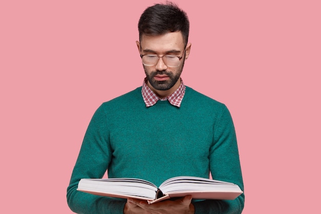 Waist up shot of serious unshaven young man gets knowledge from scientific book, wears spectacles for good vision, being diligent pupil