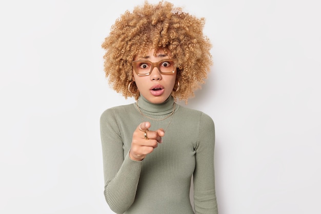 Waist up shot of shocked curly haired blonde woman holds breath from wonder cannot believe her eyes indicates forward with index finger wears casual turtleneck isolated over white background