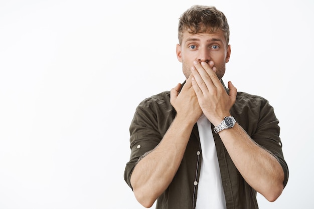 Waist-up shot of shocked speechless handsome blond guy hearing unspeakable astonishing news covering mouth with both palms and widen eyes