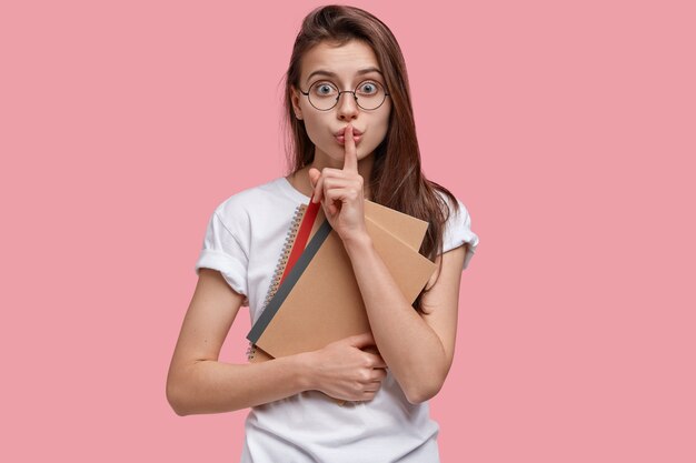 Waist up shot of shocked surprised dark haired student makes shush gesture, keeps index finger on lips, carries notepads, dressed in white t shirt