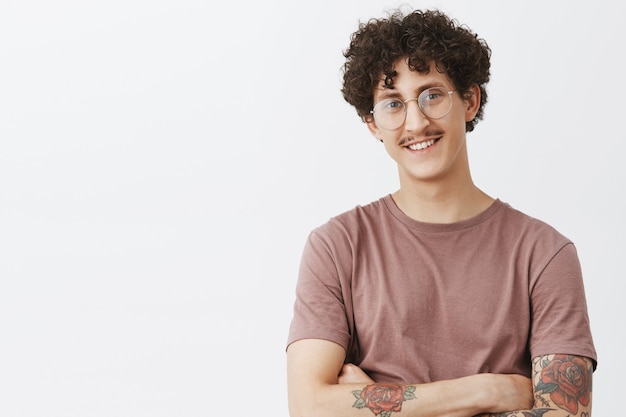 Free photo waist-up shot of stylish and creative confident happy dark-haired guy with curly hair moustache and tattooed arm smiling with self-assured satisfied expression being proud of own achievements