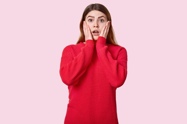 Waist up shot of surprised young woman touches cheeks, dressed in red jumper, expresses shock, poses over rosy