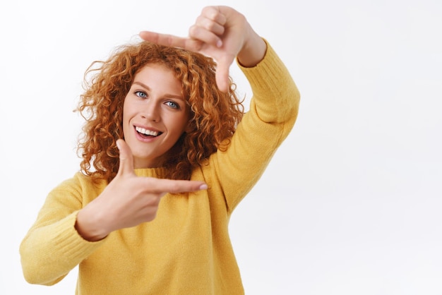 Free photo waistup portrait creative goodlooking redhead curly woman in yellow sweater photographer or designer search perfect angle look from perspective making hands frames and smiling