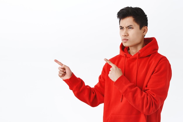 Waistup portrait of handsome asian man looking judgemental or with hate squinting frowning pointing and staring with anger and scorn left at his enemy envy standing white background