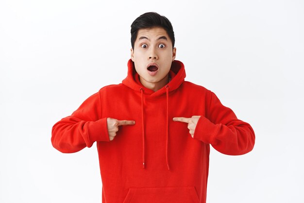 Waistup portrait of surprised amazed asian man in red hoodie shocked he was picked from all candidates pointing at himself staring camera excited volunteer standing white background