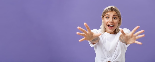 Free Photo waistup shot of amused and excited attractive stylish young woman in white tshirt pulling hands at camera with desire smiling thrilled and happy wanting hug or take something over purple background