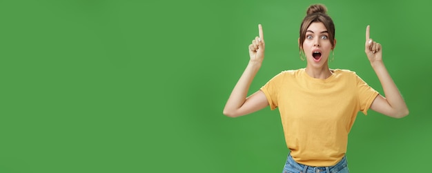 Waistup shot of interested and surprised amazed goodlooking female in yellow tshirt with tattoo dropping jaw impressed and astonished raising hands pointing up questioned against green background