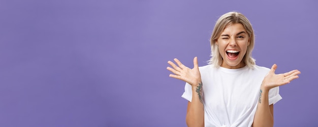 Free Photo waistup shot of playful energized sociable attractive woman with blond hair tanned skin and tattoos raising hands joyfully and spread aside winking from amusement smiling broadly over purple wall