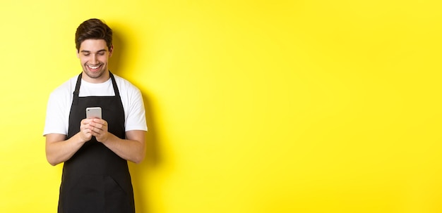 Free photo waiter in black apron reading message on mobile phone smiling happy standing over yellow background