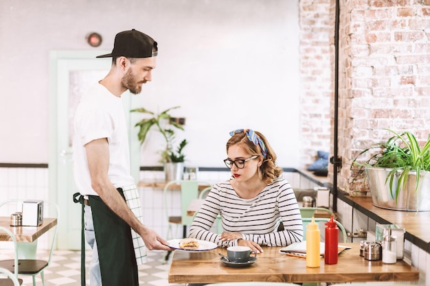 Waiter in black cap giving burger to pretty lady in eyeglasses which sitting at the table in cafe